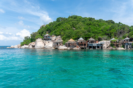 Abandoned bamboo stilt huts along a rocky tropical shoreline in Koh Tao, Thailand, with clear turquoise water and dense green vegetation.