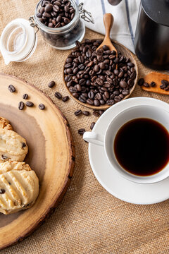 A white cup of black coffee is styled with roasted coffee beans and fresh pastries on a rustic textured tabletop.