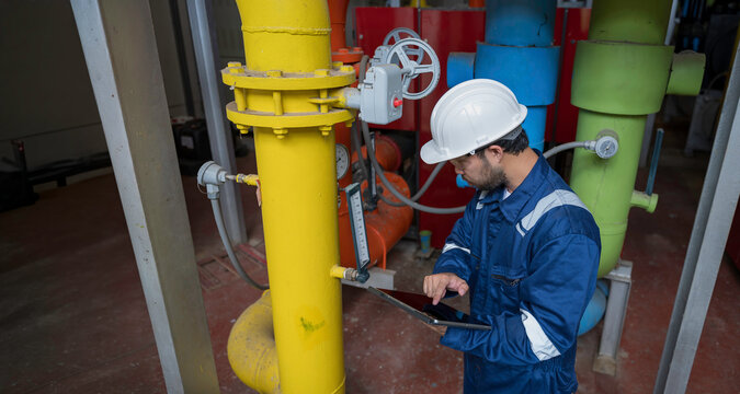 Focused professional male industrial engineer uses a laptop computer to inspect heavy machinery and colorful utility pipes during maintenance inside a modern manufacturing plant facility.