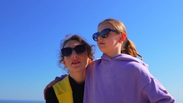 Mother and daughter laughing together on a sunlit seaside balcony under a clear sky. Static medium close-up shot.