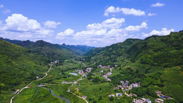 Aerial view of a green mountain valley with a rural village and winding road under a blue sky