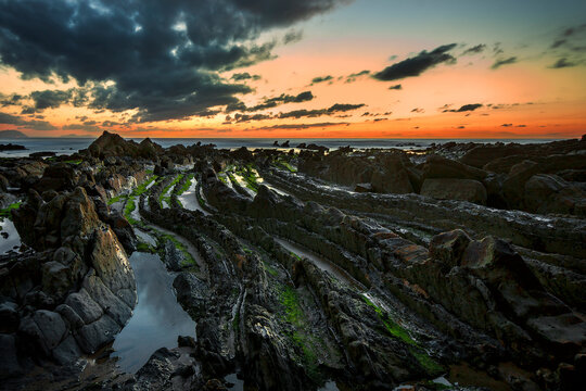 Golden sunset over the rocky flysch coastline of Barrika beach with sun reflection on wet stone platforms and ocean waves, Bizkaia, Spain