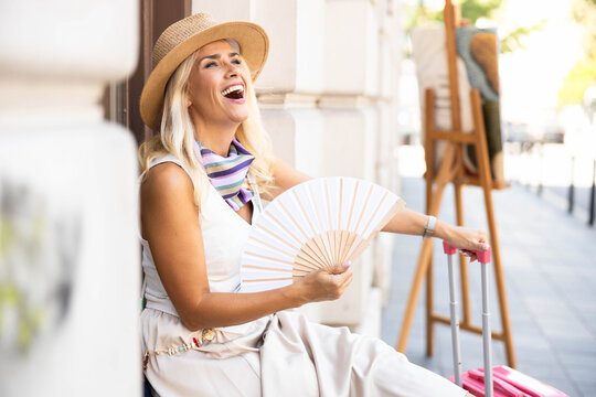 Woman traveler enjoying urban vacation, laughing cheerfully while cooling herself with a hand fan on a sunny day