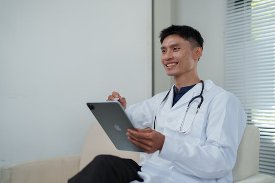 Asian doctor consulting remotely, smiling while using a digital tablet and stylus, providing telehealth services and online medical advice
