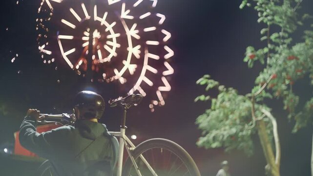 A young man in a helmet carries his bicycle on his shoulder against the backdrop of a glittering Ferris wheel at night.