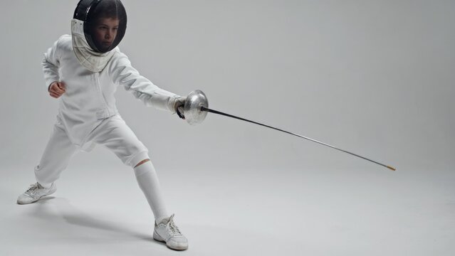 Young fencer in white uniform and protective mask holding foil, studio shot