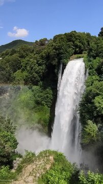 Powerful water cascade creating mist and a visible rainbow surrounded by lush green forest at Marmore Falls (Cascata delle Marmore) near Terni, Umbria, Italy