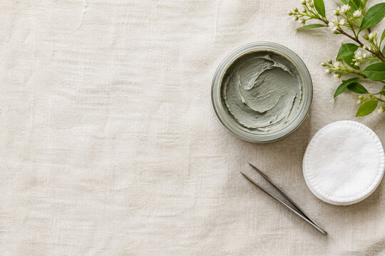 Green clay face mask in a glass jar with cotton pad and tweezers on linen