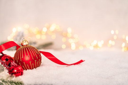 Red and gold Christmas baubles with ribbon resting on snow, surrounded by warm fairy lights in a cozy, festive holiday setting. Ideal for holiday greeting cards, festive banners.