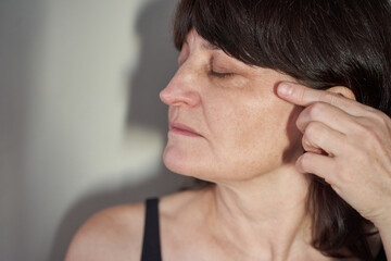 Middle-Aged Woman Pressing Temple With Fingertip While Eyes Closed In Indoor Profile Portrait Against Neutral Wall Showing Facial Tension And Attempted Relaxation In Soft Light. © elenavolf