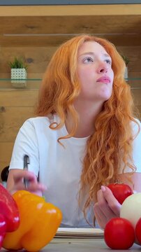 Thoughtful young woman with vibrant red hair meticulously plans her healthy meal, writing down fresh ingredients and recipes for nutritious, balanced eating and well-being.
