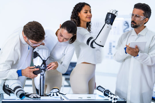 Robotics prototype evaluation, two heads dip together over the articulated robot arm on its stand while the wearable limb stays lifted clear of everyone's shoulders