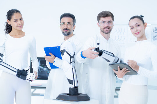 Bionic engineering workshop: two hands close around the black five-finger gripper of a lab robot arm as the wider team looks on from behind the illuminated white plinth