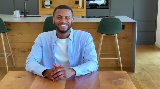 Joyful young Black man waves and smiles during a vibrant video call on his laptop from a modern kitchen setting, perfectly embodying remote communication and digital connection.