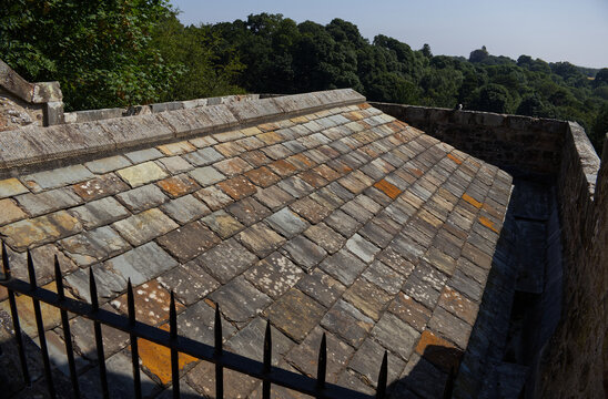 Traditional weathered stone slate tile roof on a historic building enclosed by a parapet wall, overlooking green summer trees.
