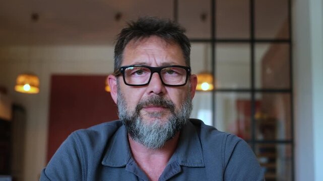 A bearded man with glasses is engaged in a thoughtful conversation, looking up and then making a discreet "shhh" gesture. He conveys a sense of seriousness and careful communication.