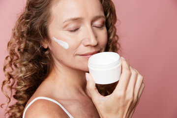 Middle-aged woman with natural skin holding cream jar on pink background. Authentic beauty and...