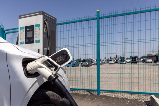 White electric car is parked at a modern charging station with a cable plugged in. Clear blue sky and a parking lot filled with vehicles create a vibrant outdoor scene.
