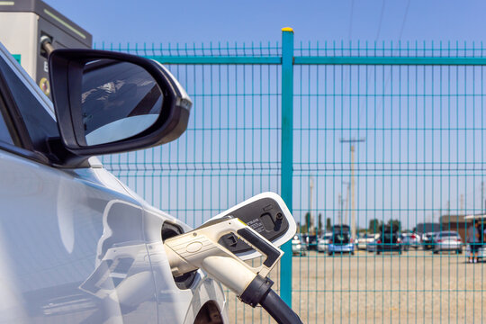 Close-up view of electric vehicle charging at outdoor station. Charging plug is inserted into the vehicle's port, with a turquoise fence and parking lot in the background.
