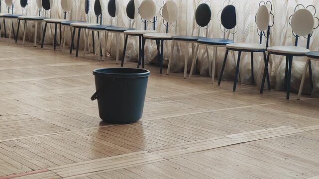 Empty bucket sitting on a wooden parquet floor to collect water from a leaking ceiling. Damaged room with chairs waiting for repair