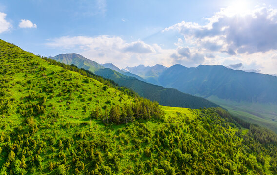 Aerial view captures vibrant green hills with dense vegetation leading to majestic mountains in Kyrgyzstan. Sunlight filters through clouds, enhancing the serene landscape.