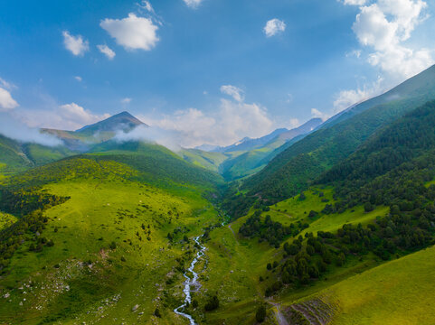 Aerial perspective reveals a breathtaking mountain valley bathed in daylight. A clear, shallow river snakes through the vibrant green landscape, flanked by rolling hills and steep mountain slopes.