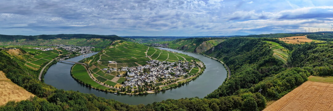 Panoramablick auf die Moselschleife bei Minheim mit idyllischen Weinbergen, blauem Flusslauf und typischem Weindorf unter leicht bew&ouml;lktem Sommerhimmel.