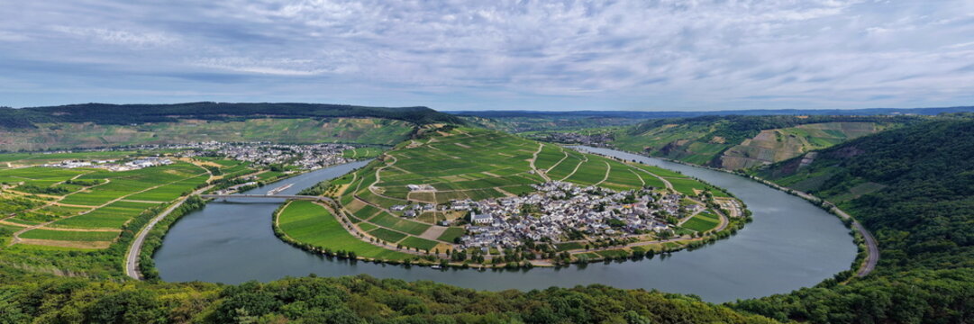 Panoramablick auf die Moselschleife bei Minheim mit idyllischen Weinbergen, blauem Flusslauf und typischem Weindorf unter leicht bew&ouml;lktem Sommerhimmel.