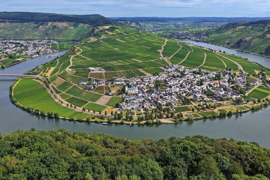 Panoramablick auf die Moselschleife bei Minheim mit idyllischen Weinbergen, blauem Flusslauf und typischem Weindorf unter leicht bew&ouml;lktem Sommerhimmel.
