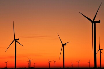 Obraz premium Vue panoramique d'un champ d'éoliennes dessinant des silhouettes verticales au coucher du soleil, sous un ciel orange mauve. Symbole d'une énergie renouvelable grâce à la force du vent.
