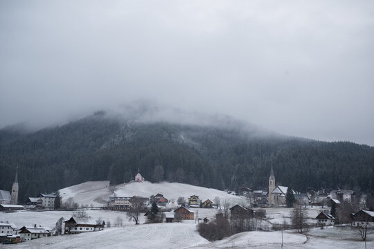 Wide shot of misty alpine village in Gosau, Austria, with church spires and snow covered fields. Pine forest covered mountain peaks shrouded in low fog on cold winter day.