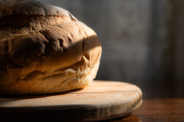 A rustic round loaf of bread sits on a wooden board in dim lighting. Concept of artisanal baking, homemade food, and traditional culinary arts. © kinomaster