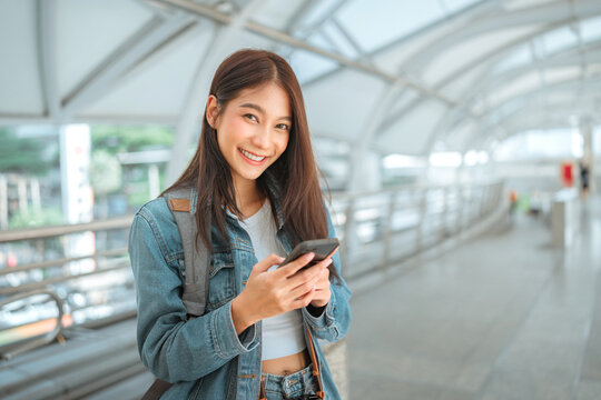 Smiling young woman with backpack using smartphone on modern covered walkway, enjoying mobile technology, digital communication and online connection while commuting.