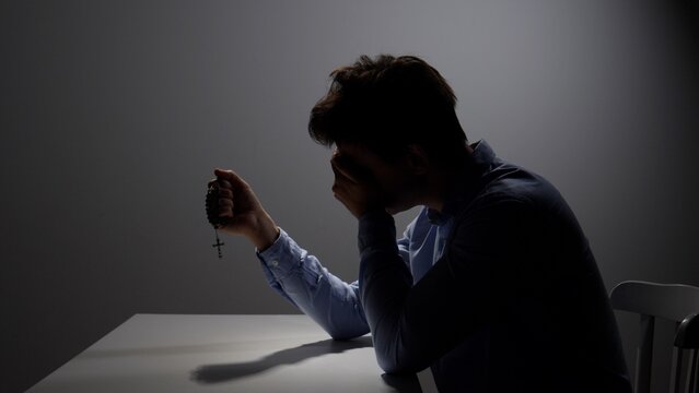 Man sits alone at a dim table, covering his face while clutching rosary beads in a moment of deep grief, prayer and anguished solitude seeking comfort and faith