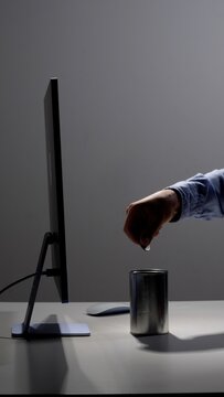 Person's hand dropping a single coin into a metal tin can, representing concepts of digital finance, online donation, savings, and charitable giving while working at a computer desk