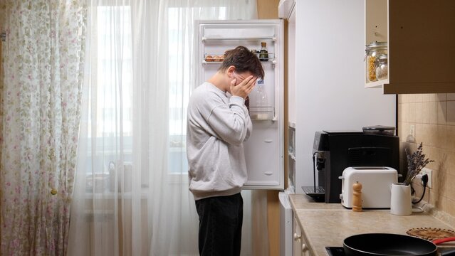 Young man covering his face with hands, feeling stressed and desperate while looking into a nearly empty refrigerator, representing hunger, food insecurity, and financial struggle