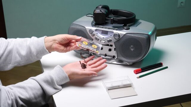Person's hands holding a broken cassette tape with tangled magnetic tape, ready to fix it with a pencil, while a vintage boombox and headphones rest on a white table