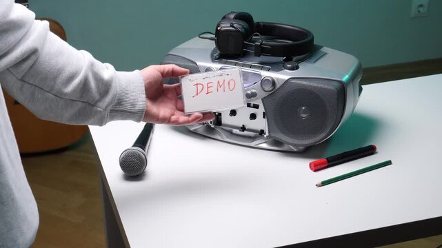 Person's hand holding a cassette tape labeled demo near a boombox, microphone, and headphones, symbolizing the process of recording music, creating a demo, or recalling vintage audio technology