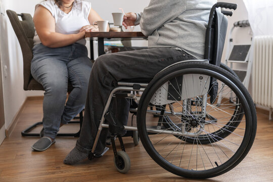 Person with disability and his personal assistant are in a kitchen drinking coffee and tea while talking.