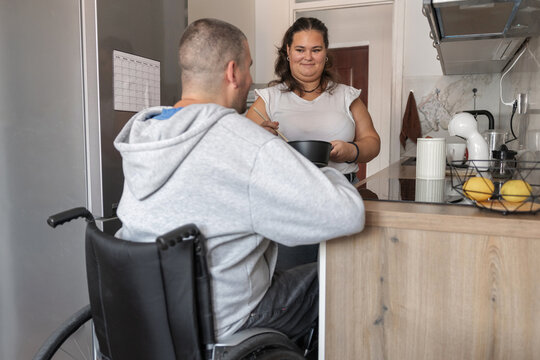 Female personal assistant helps a person in a wheelchair with meal preparation in a kitchen. They work together for independent living.