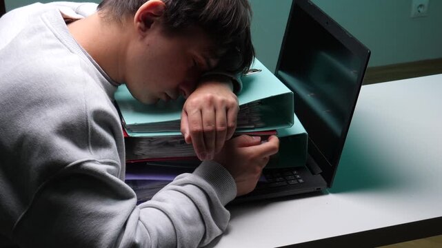 Young man sleeping on a stack of binders and documents next to a laptop, representing exhaustion, burnout, and overwhelming workload from studying or working late