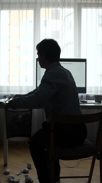 Vertical video silhouette of a male freelancer at a desk, hunched before a laptop amid crumpled paper, conveying creative block, stress and burnout during a long workday