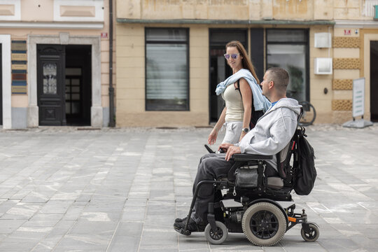 A person using a wheelchair is assisted by woman personal assistant in a city. They are engaged in a conversation while exploring the area.
