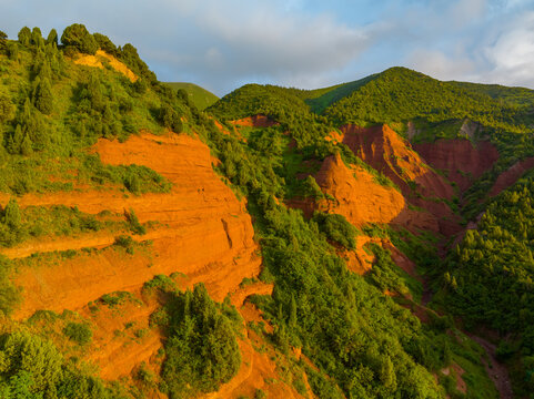 Aerial view captures a rugged mountain landscape illuminated by golden hour light in Kyrgyzstan. Striking orange-red rock formations contrast with lush green vegetation, creating a serene and majestic