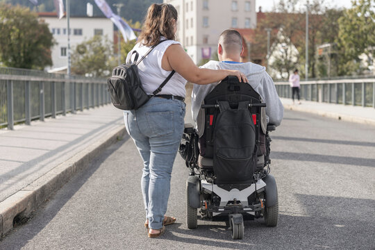 A female personal assistant assists a male in a wheelchair on a city street during daylight. They navigate together with confidence and support.