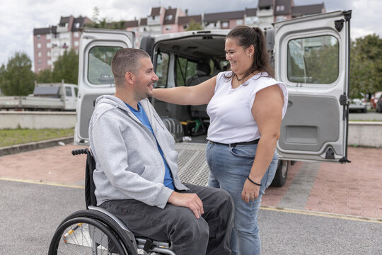 A person with a disability is getting help from a female personal assistant by an adapted vehicle. They are in a parking lot.