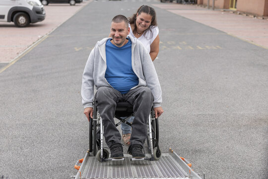 A man in a wheelchair is assisted by a female personal assistant as they use a ramp for accessible transportation. They are in an open parking area.