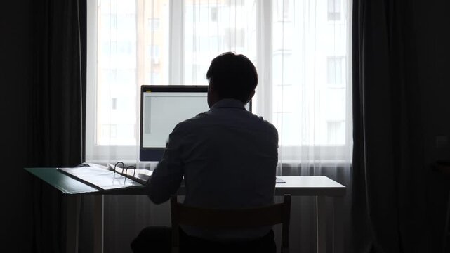 Businessman silhouette in a home office, clutching his head in frustration at a computer, backlit by a bright window during a stressful remote workday
