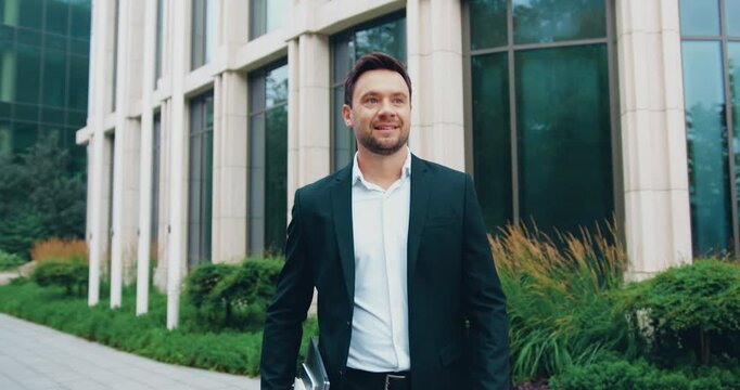 Successful businessman in a formal suit walking through a modern urban park near a contemporary glass office building.
