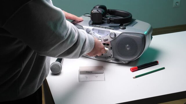 Person's hands adjusting controls on a silver boombox with headphones, a microphone, and a cassette labeled punk on a white table, engaging in vintage audio recording